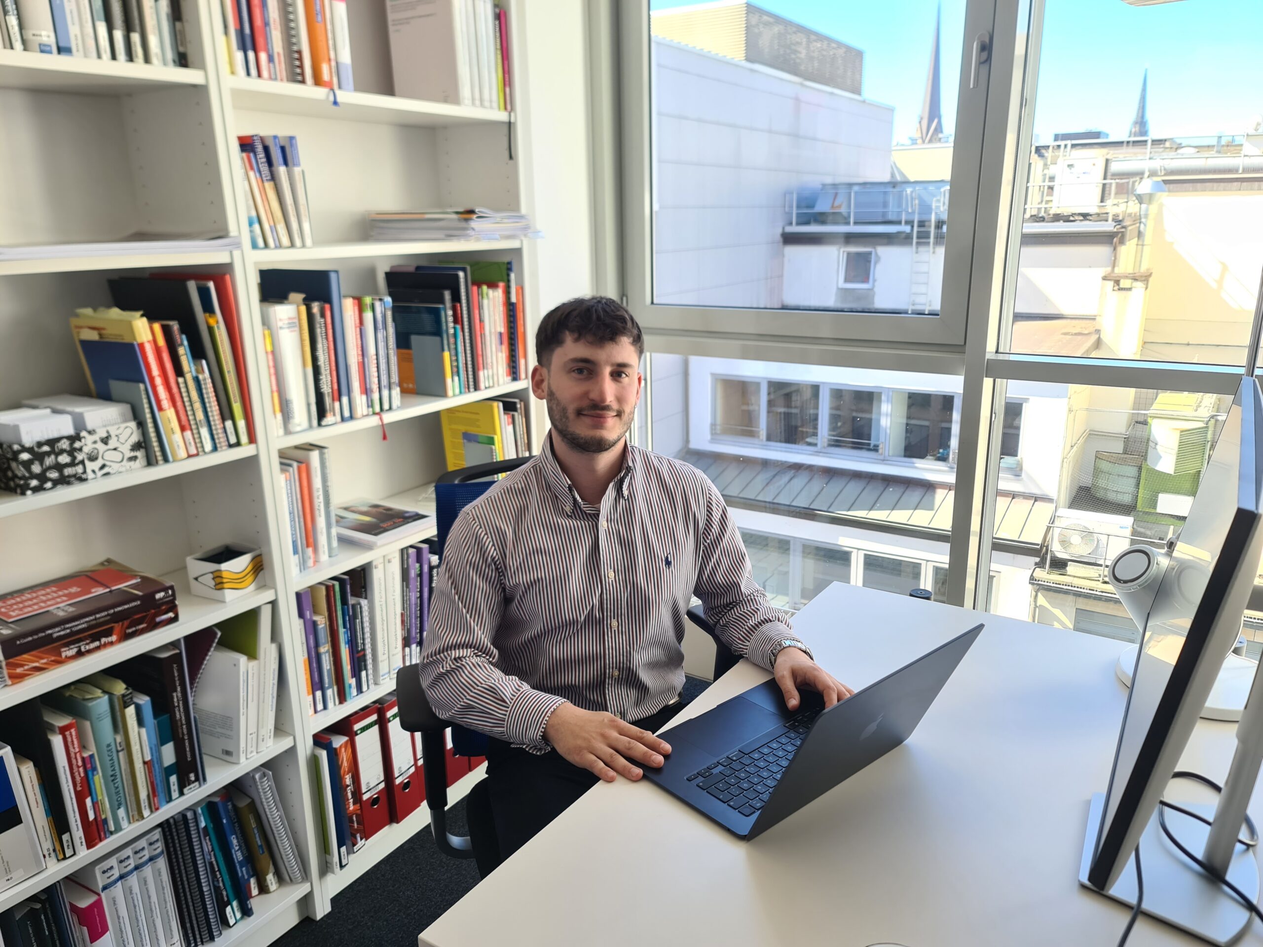 Dominik Marzec von House of PM sitzt an einem Schreibtisch in einem hellen Büro mit Bücherregal.